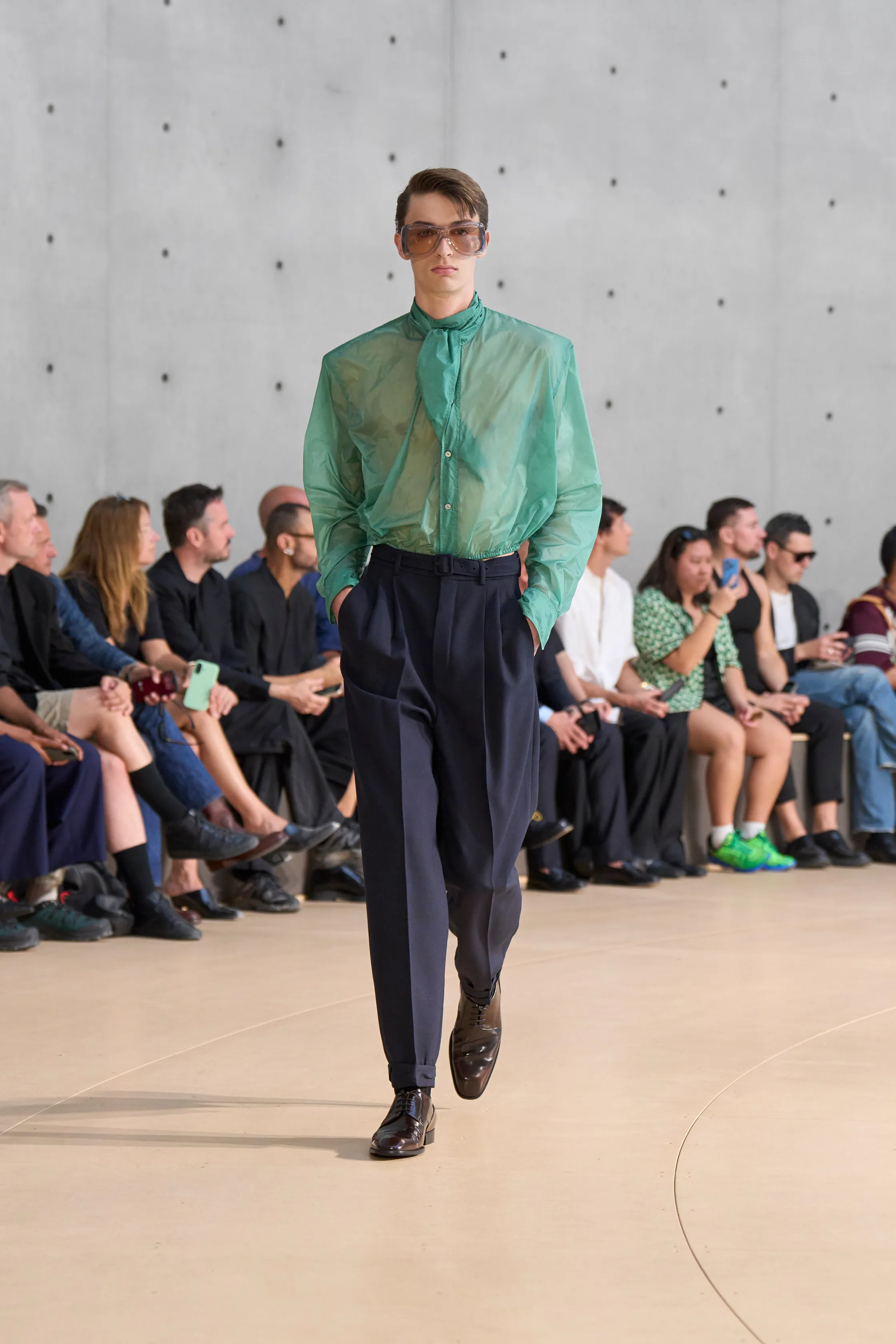 A model wears a translucent mint green shirt with a tied collar, tucked into high-waisted navy trousers. The look is styled with gradient sunglasses and glossy black shoes.