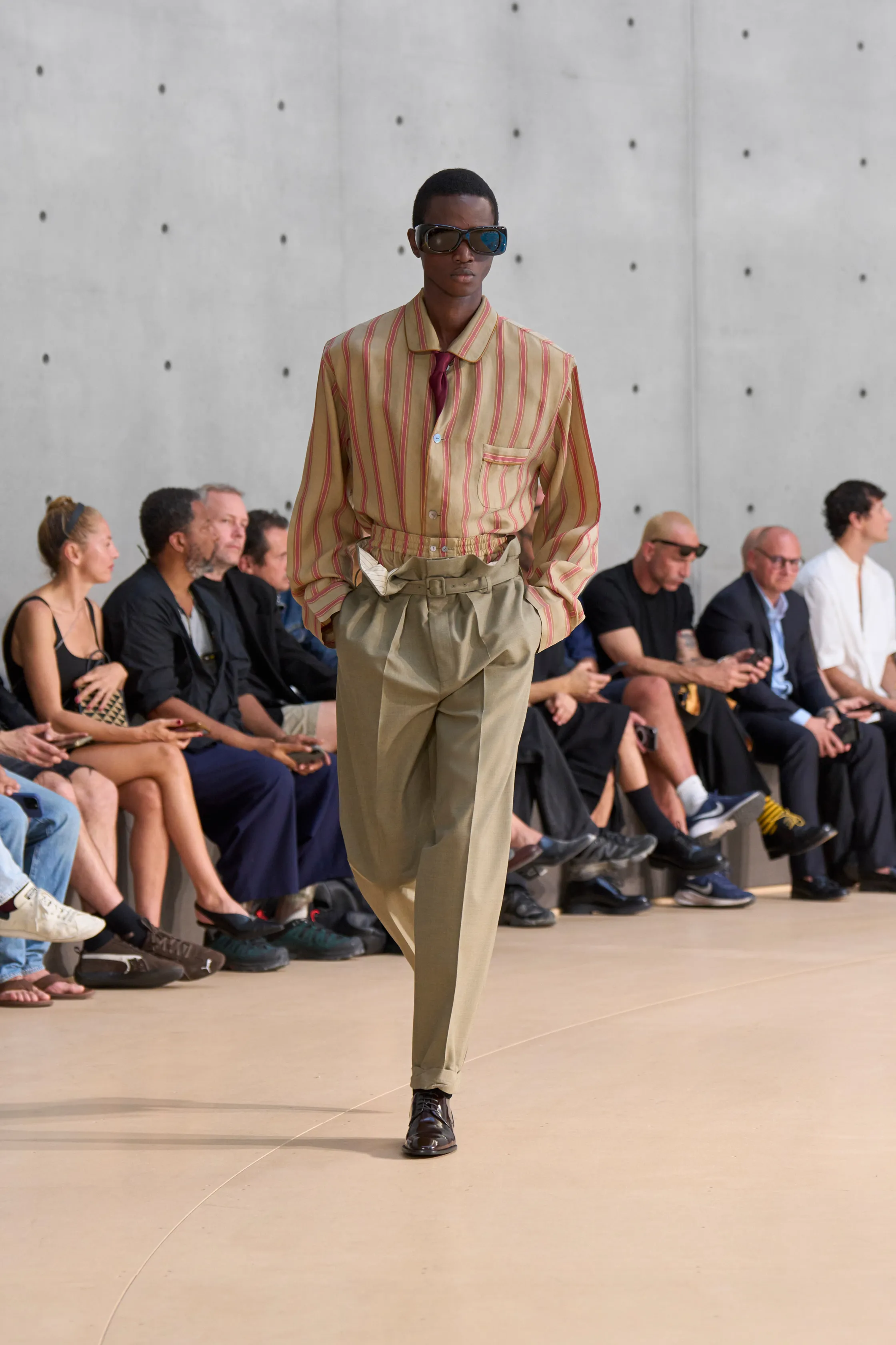 A model walks the runway wearing a beige and red striped button-up shirt tucked into high-waisted pleated khaki trousers, cinched with a wide belt. He pairs the look with oversized sunglasses and dark leather shoes.