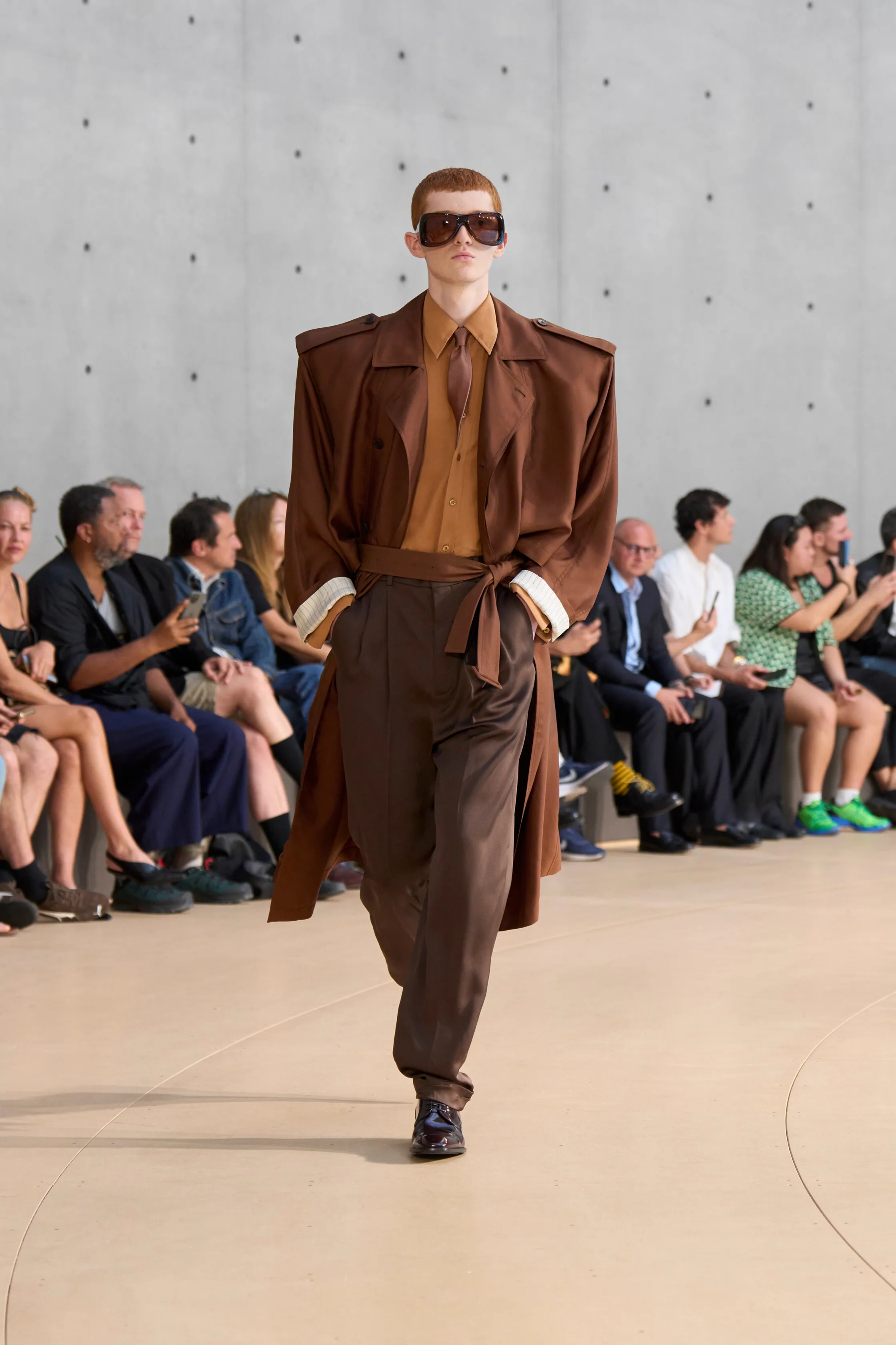 A red-haired model wears a chocolate brown trench coat over a golden brown shirt and tie, matched with wide-legged brown trousers. The look is finished with large square sunglasses and black shoes.