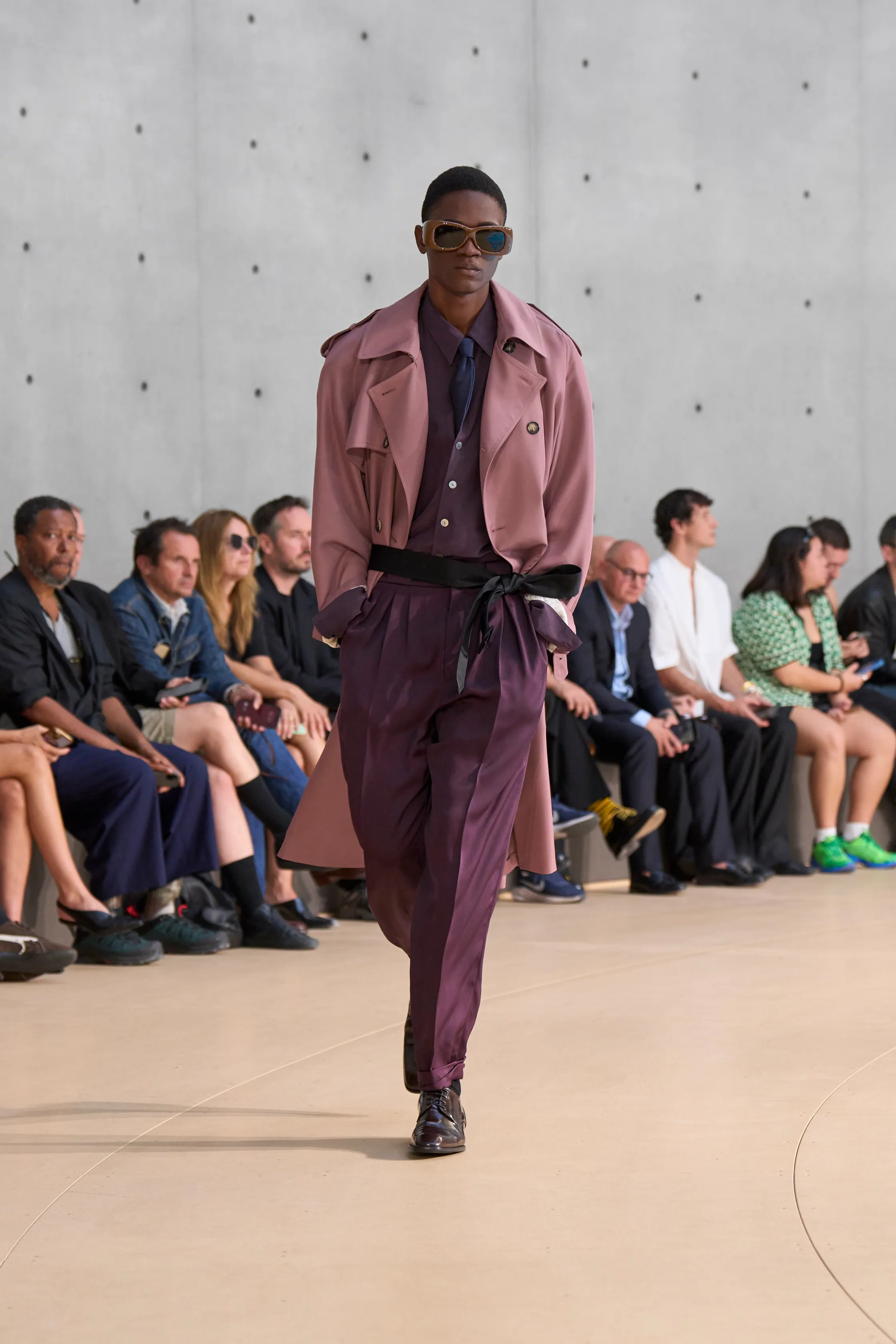A model showcases a dusty pink trench coat over a deep plum-toned ensemble, including a shirt, tie, and wide trousers. The look is belted at the waist and styled with gold-rimmed sunglasses.