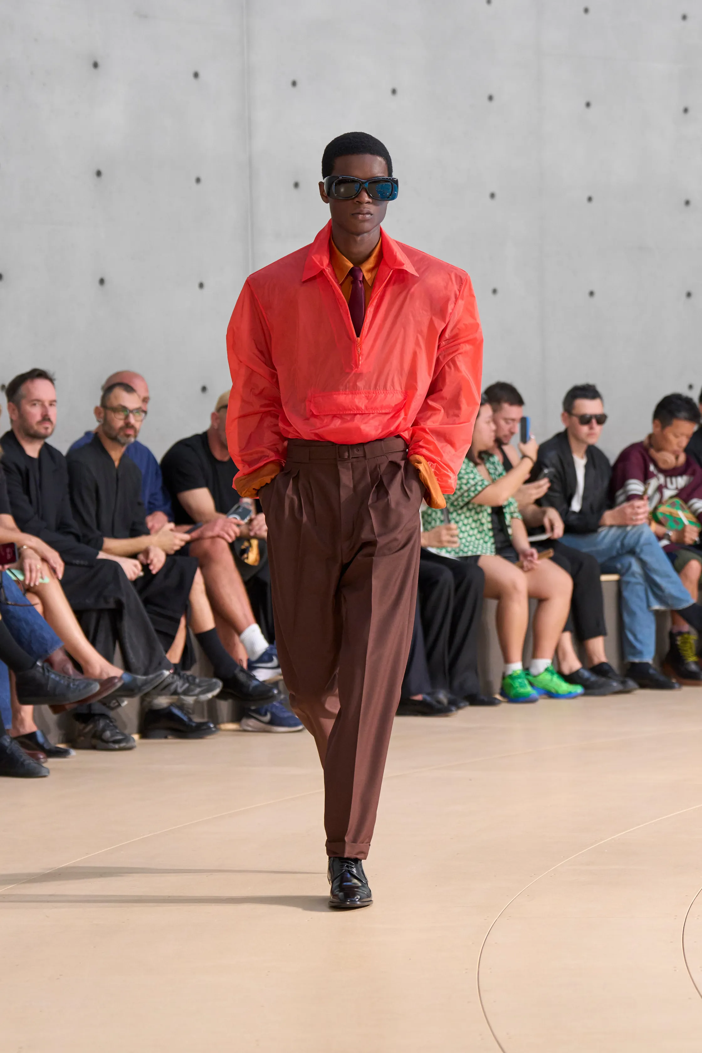 A model sports a vivid crimson windbreaker over an ochre shirt and burgundy tie, paired with chocolate brown trousers. Oversized black sunglasses and sleek shoes complete the bold palette.