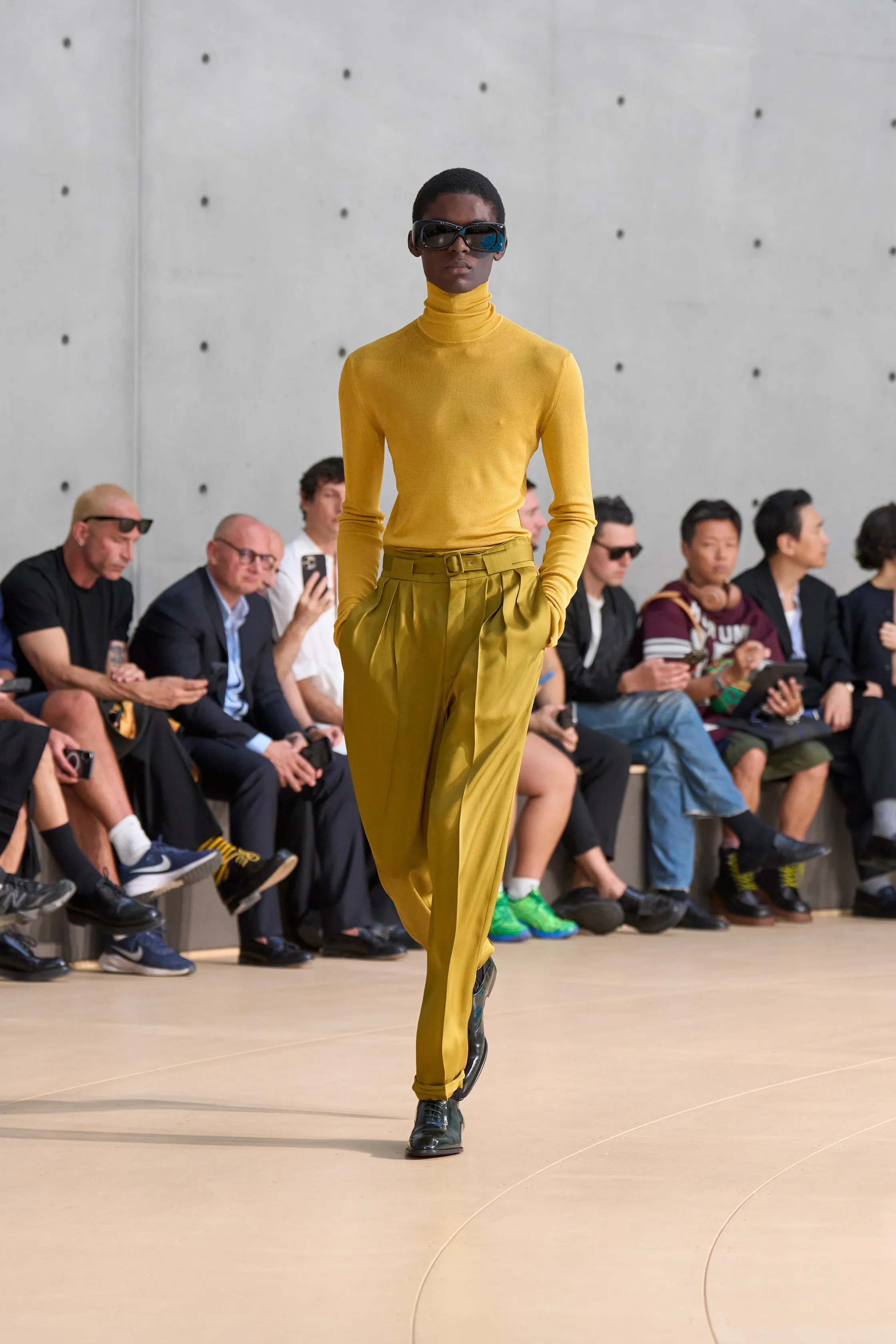 A model wears a fitted mustard yellow turtleneck tucked into matching pleated trousers. The clean silhouette is accented by dark sunglasses and glossy black shoes.