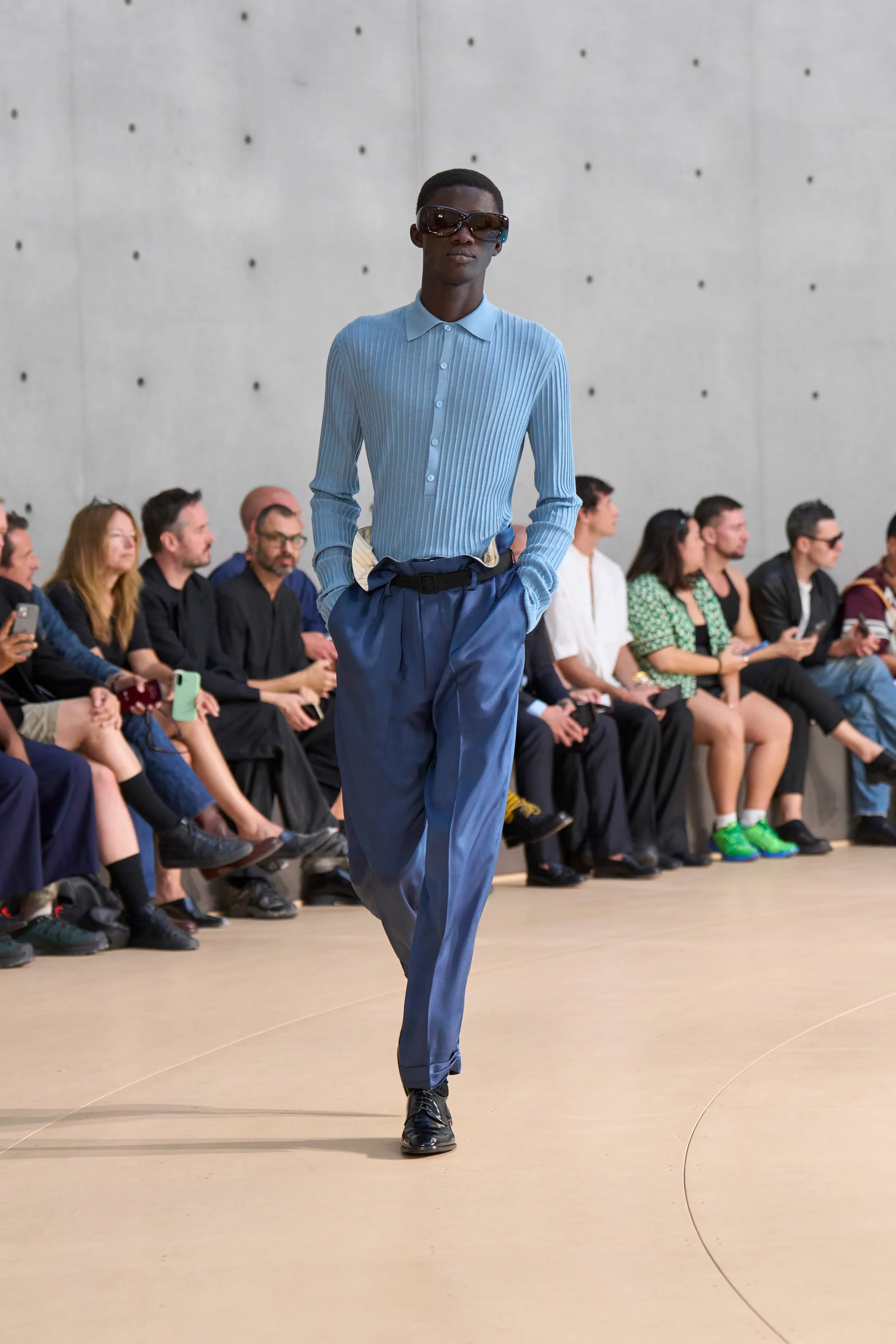 A model walks in a pale blue ribbed polo-style shirt with long sleeves, tucked into silky cobalt trousers. The look is finished with a black belt and dark sunglasses.
