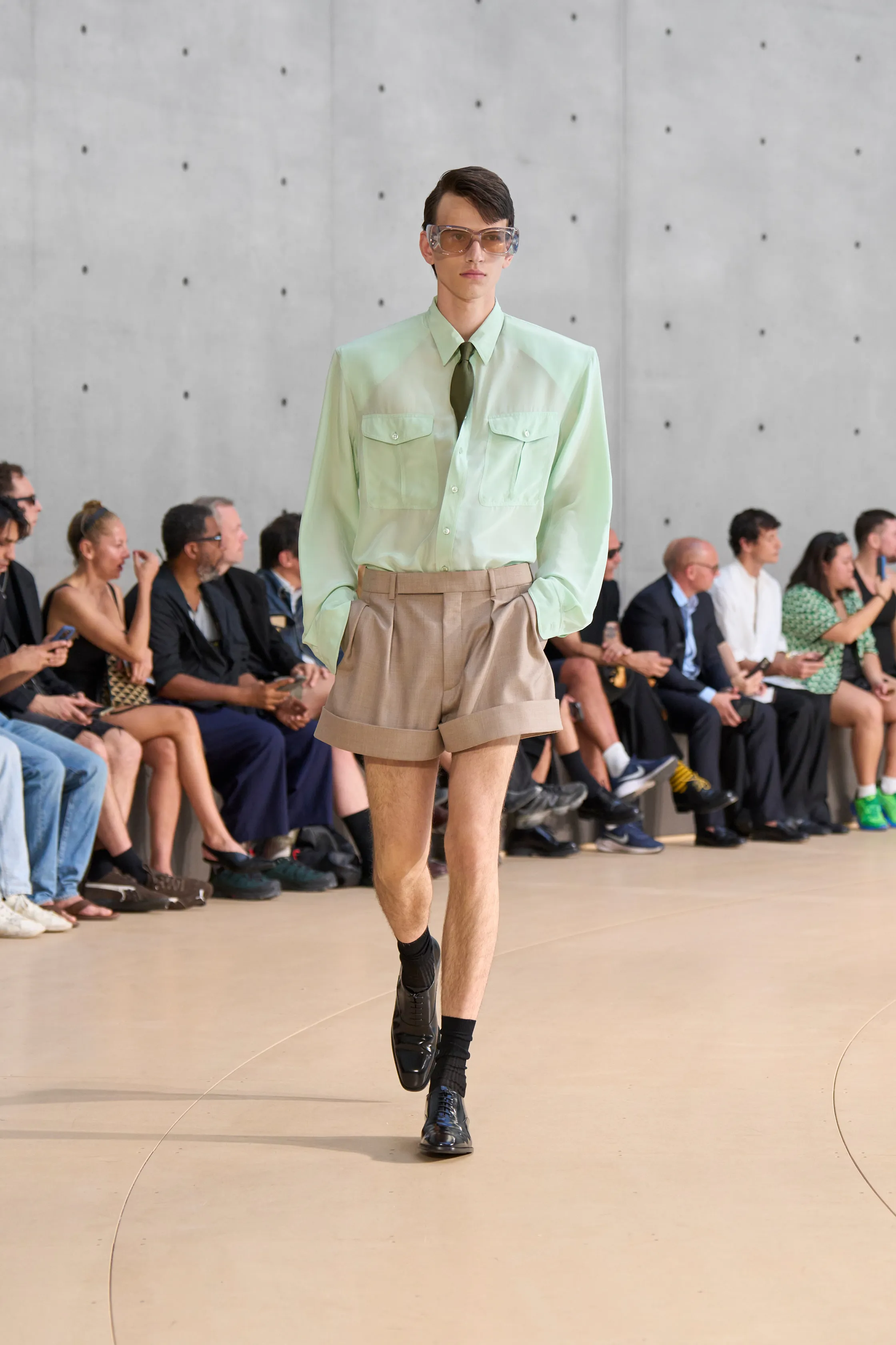 A model wears a crisp mint green dress shirt with two chest pockets and a tie, tucked into tailored tan shorts with thick cuffs. The outfit is accessorized with oversized clear sunglasses and dress shoes with socks.