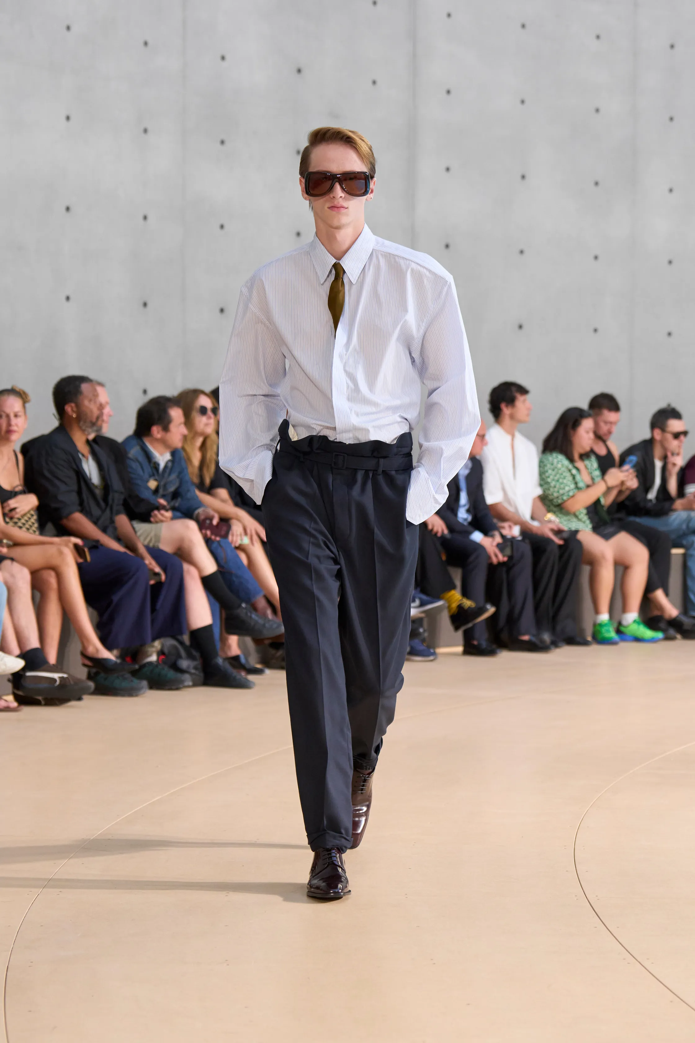 A model presents a classic white pinstriped shirt with a gold tie, paired with navy high-waisted trousers. Oversized brown sunglasses and glossy black shoes complete the clean, minimal look.
